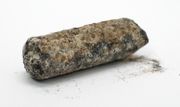 A heavily pitted, blackish cylindrical rod, with extensive, crumbling brown-and-white corrosion
