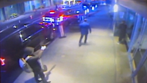 A dirty frame of CCTV footage covering the sidewalk. The killer stands at the bottom left, wearing a hooded jacket, training his sidearm at Johnson, in the center, his back facing the camera.