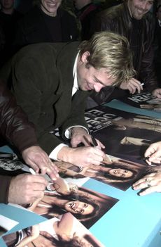 A Caucasian male bent over a table autographing a movie poster. He has light brown hair with blonde highlights, and is wearing a dark-colored trench coat with a white shirt. Visible in the background and foreground are other people, some of whom are also signing autographs.
