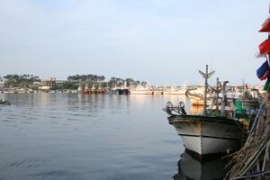Two boats anchor in a harbor on the right. Blue skies and the sea are clear and tranquil.