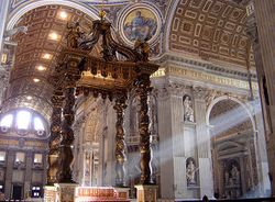 Photo shows the baldachin standing in the centre of the church, viewed looking towards the nave. There is an altar beneath it which has a red and gold frontal cloth decorated with large crosses.