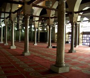 A large room filled with rows of cylindrical columns on top of square bases. The columns support arches which are pierced by square beams going the length of the room in both directions. Hanging from the beams are lamps, and the room's floor is covered with a red carpet with a repeated beige arched doorway shaped design on it. Exterior light enters from right of the room.
