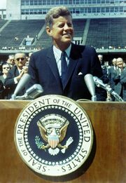 President Kennedy is pictured speaking behind a podium. Rice University's stadium is visible behind him.
