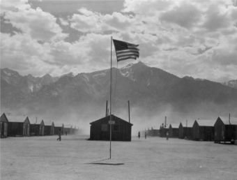 Small buildings in rows in a dusty flat area. A tattered U.S. flag flies from a pole in the foreground, and tall mountains dominate the background below a sky with clouds.