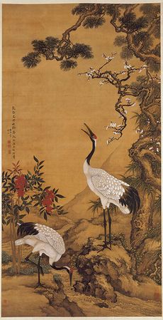 Two cranes near a pine tree. One is feeding on the ground while another rears its head high. Red flowers are also in the background