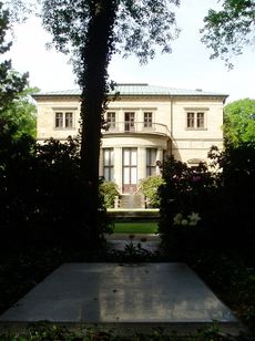 A grey slab, level with the ground framed by bushes and in the shade of the tree. In the background a fountain and a large two-storied house with a balcony.