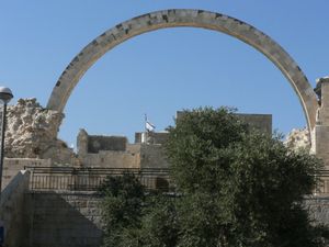 A solitary stone arch rising into the blue sky, spans across one of the former sides of the building. A tree, with thick green foliage is seen in the foreground.