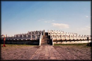 Color photograph of a three-level stone structure with railings on each level, viewed from the outside, facing a staircase that leads to the top level.