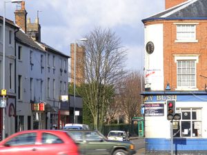 The corner of a street with a public house called The Ivy Bush on the left side. In the background two tall brick towers can be seen further left.