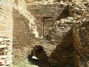 Inside daytime view of a ruined and ceiling-less rectangular room. Tawny-beige stacked sandstone bricks compose walls rising from brush-covered ground. The several walls visible in the image are up to perhaps a dozen feet in height. In the wall immediately at center, a triangle-shaped entrance several feet high leads to an adjacent chamber behind. The upper part of the same wall, shaped like an inverted-triangle, has fallen away or otherwise been removed, revealing a rectangular doorway leading to yet another concealed room. At left and right are two similar walls perpendicular to the one at middle.