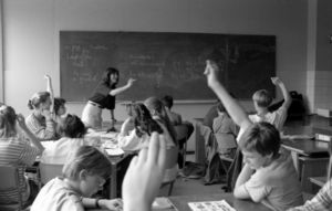 Students are seated at tables in a classroom. Some students have their hands raised and a teacher is pointing to one of these students.
