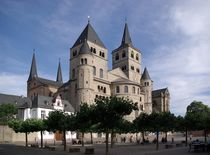 A huge cathedral with numerous towers, both square and round, rises above a town square where people are sitting in the shade of clipped trees.