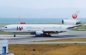 A McDonnell Douglas DC-10 aircraft taxiing on the tarmac, with a yellow grass strip in the foreground and a mountain and seaview in the background