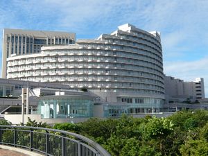 large circular multi-story building with stepped upper levels, facade comprised of multiple rows of balconies; foreground is a tree-lined park area, with a curved walkway railing, and additional buildings behind.