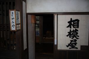 Dimly-lit narrow shopfront, with large black characters written in a single column on a shoji. A washi-and-frame lantern, mounted to protrude from the pillar, also bears signage.