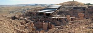 Roughly built stone walls surrounding T-shaped stone pillars under a modern steel walkway and roof in a hilly landscape