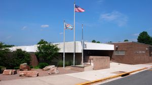 An entrance to a school below a white concrete overhang with red brick walls. National and local flags fly next to a circle of five rocks.